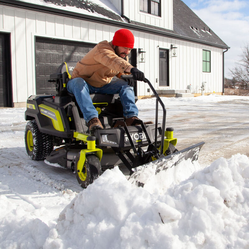 A person wearing a red beanie and casual clothes operates a green and black snow plow vehicle on a driveway of a house with black garage doors. The vehicle pushes a pile of snow in a snowy outdoor setting.