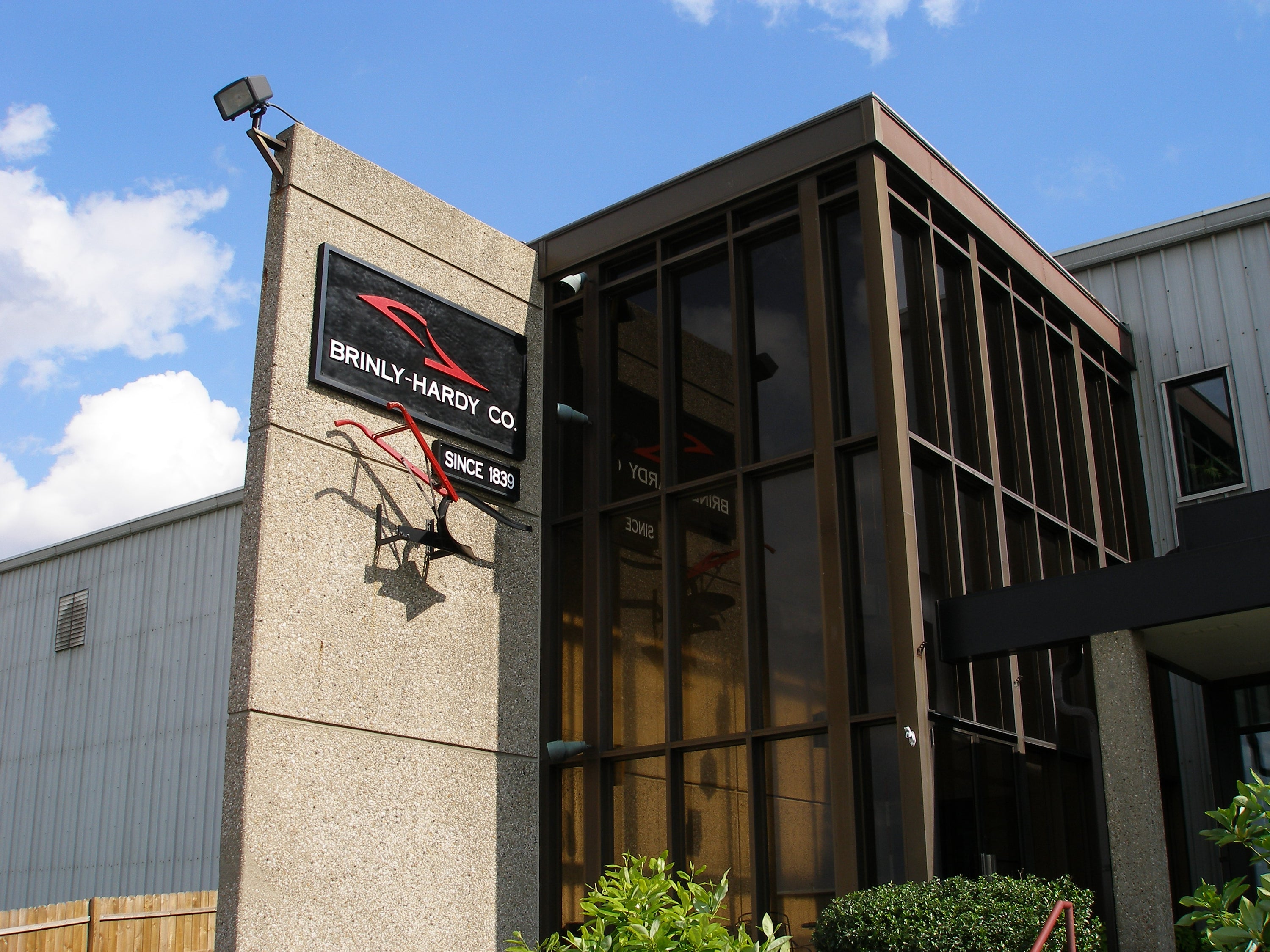 Industrial building with a modern glass entrance and concrete exterior. A sign reads BRINLY-HARDY CO., Since 1839. The area is surrounded by greenery under a blue sky with clouds.