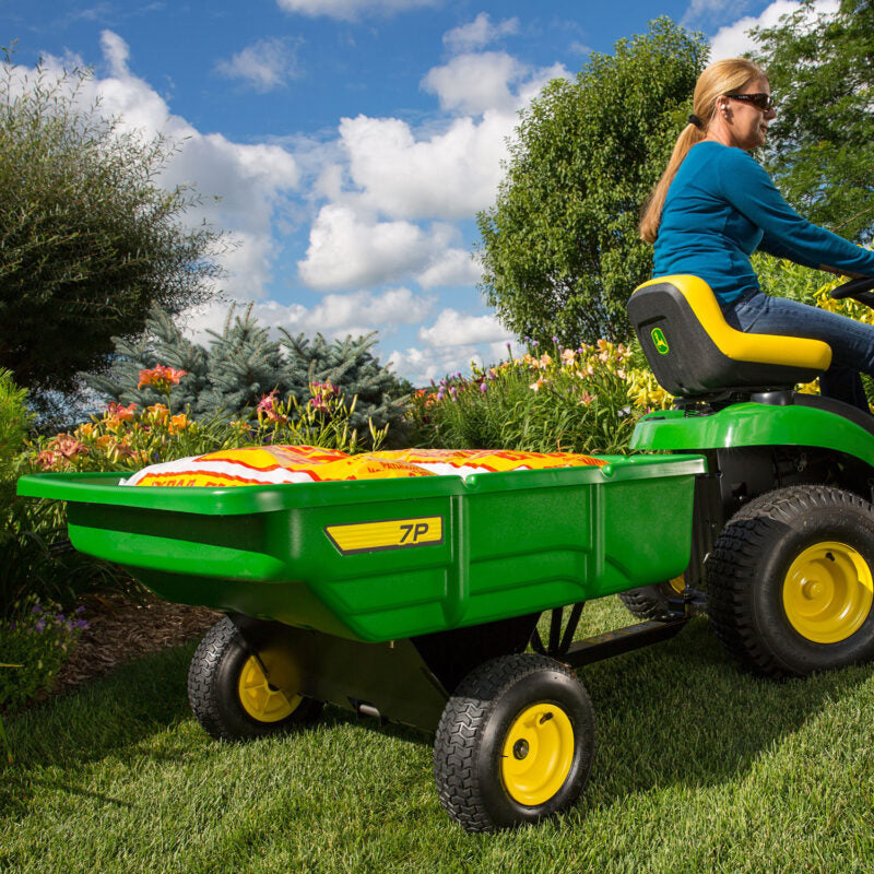 A person wearing a blue sweater operates a green and yellow ride-on lawnmower with a 7P model trailer attached. The lawnmower is on a grassy area surrounded by lush greenery and colorful flowers under a partly cloudy sky.
