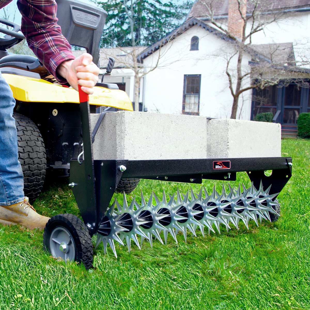 A person uses the Brinly Parts 40 Tow-Behind Spike Aerator, model SAT2-40BH-P, with a spiked roller and cinder blocks for weight, hitched to a yellow lawn tractor. On lush grass before a house with trees, this setup improves soil penetration as they wear jeans and brown boots.