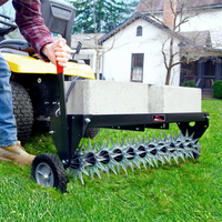A person uses the Brinly Parts 40 Tow-Behind Spike Aerator, model SAT2-40BH-P, with a spiked roller and cinder blocks for weight, hitched to a yellow lawn tractor. On lush grass before a house with trees, this setup improves soil penetration as they wear jeans and brown boots.