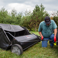 A person in a green shirt and red gloves, wearing a hat, kneels on grass adjusting a Brinly Parts 54″ Lawn Sweeper with Double-Helix Brushes (LS2-54BH-G) under an overcast sky with pine trees in the background, focusing intently on ensuring all components are perfect.
