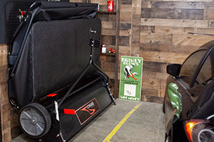 Folded treadmill stored vertically in a garage next to a parked car. The wooden wall has a poster featuring a person stepping on a rake with the text Careful Rakes and Step Wisely. A small fire extinguisher is mounted above.