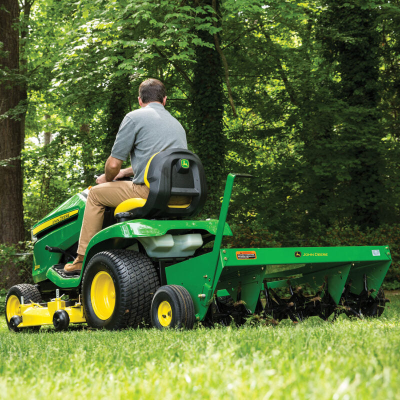 A person wearing a gray shirt and brown pants operates a green John Deere lawn tractor with an aerator attachment in a lush, green garden surrounded by tall trees.