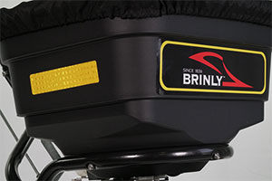 Close-up of a black Brinly spreader with a yellow label on the side. The Brinly logo and text Since 1839 are visible on a black and red panel. The spreader appears to be part of lawn or agricultural equipment.