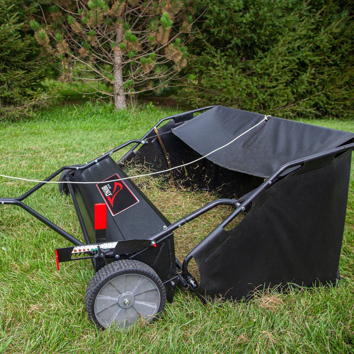The Brinly Parts 54″ Lawn Sweeper with Double-Helix Brushes (LS2-54BH-G) features two large gray wheels and a partially filled collection bag. It rests on green grass, complete with a handle for easy maneuvering, against fir trees.