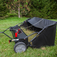 The Brinly Parts 54″ Lawn Sweeper with Double-Helix Brushes (LS2-54BH-G) features two large gray wheels and a partially filled collection bag. It rests on green grass, complete with a handle for easy maneuvering, against fir trees.