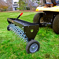 A Brinly Parts 40 Tow-Behind Spike Aerator (SAT2-40BH-P) with a double tow bar is attached to a yellow riding lawn mower, expertly perforating the soil for healthier roots. Houses and trees frame the lush yard.