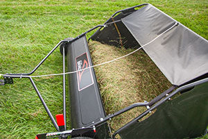 A lawn sweeper filled with grass clippings is positioned on a grassy area. The sweeper has a black collection bag and a red and black frame, with a handle visible for pushing or towing.