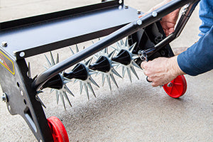 A person adjusts a black metal aerator tool with red wheels on a concrete surface. The tool features multiple spikes for aerating soil. The person is wearing a blue long-sleeve shirt.