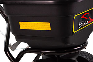 Close-up of a black Brinly spreader hopper with a yellow label on its side. The hopper is mounted on a metal frame and has a visible tire on the right side.