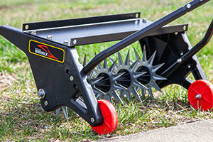 A manual lawn aerator with spiked wheels and a black frame rests on grassy ground. The red wheels and metal spikes are prominently visible, designed to puncture the soil to improve lawn health.