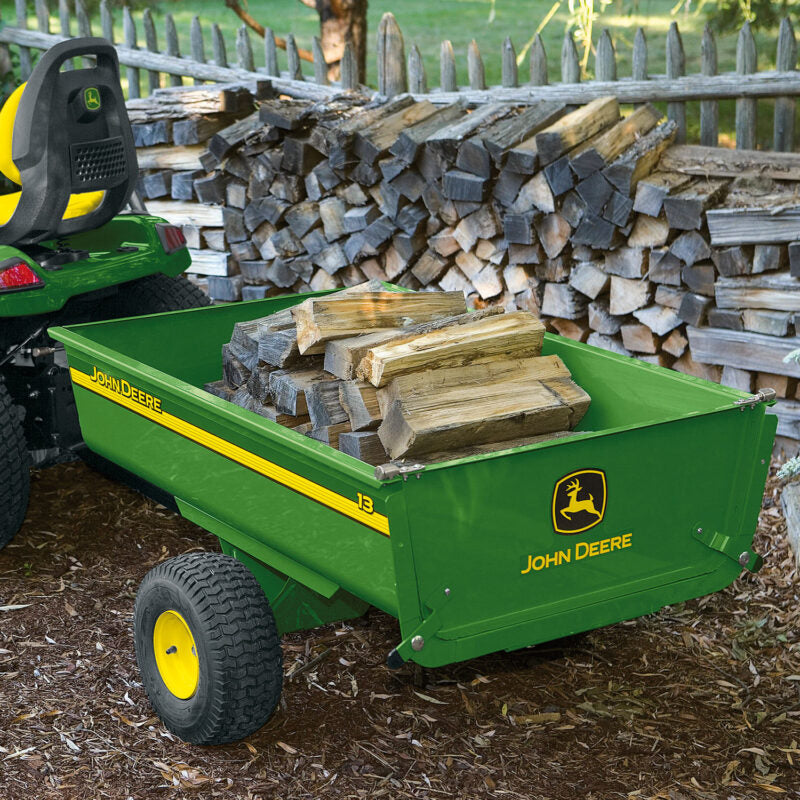 A green John Deere trailer loaded with firewood is attached to a lawn tractor. It is parked on a mulch-covered ground, with neatly stacked firewood and a wooden fence in the background.
