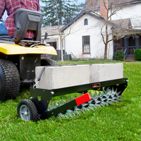 A person on a yellow lawn tractor uses a Brinly Parts 40 Tow-Behind Spike Aerator with Extra-Strength Double Tow Bar & Transport Wheels (SAT2-40BH-P) laden with concrete blocks to aerate the grassy lawn, with a white house and trees in the background.