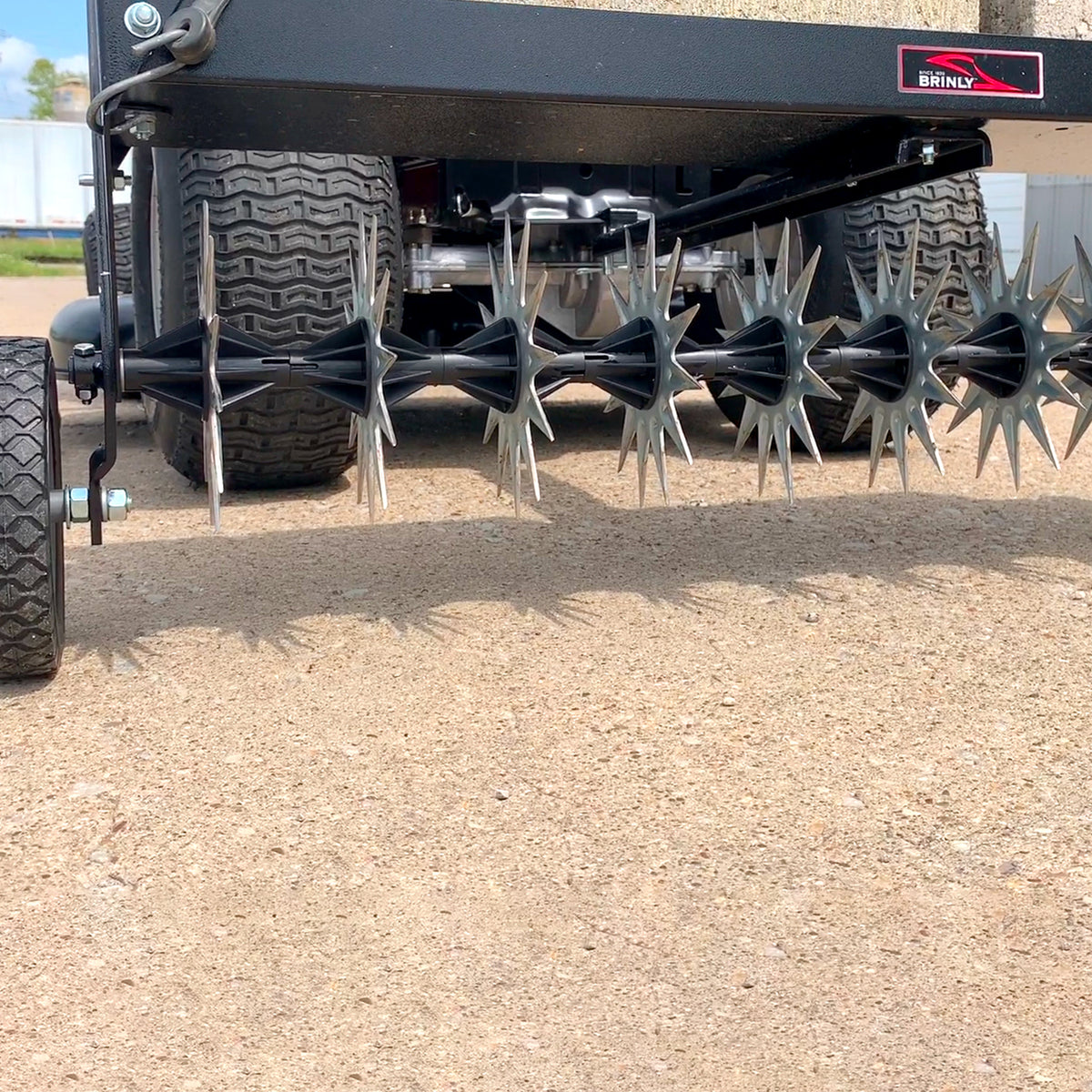 Close-up view of the Brinly Parts 40 Tow-Behind Spike Aerator (SAT2-40BH-P) on concrete. The spiked wheels enhance soil penetration for better airflow and nutrient absorption, promoting a healthier lawn root system. It features an extra-strength double tow bar and is attached to a black frame.