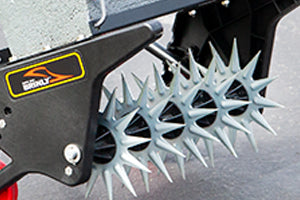 Close-up of a spike aeration tool with sharp, star-shaped metal spikes attached to a black frame. The spikes are designed to puncture the ground, promoting soil aeration. The surface below is a textured, gray concrete.