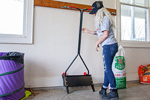 A person with long hair and a cap is assembling a gardening tool in a garage. A bag of lawn food and other supplies are nearby. The person is adjusting the handle of a tool with red wheels. They are wearing jeans and boots.