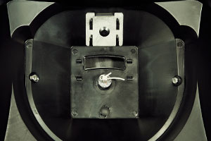 Close-up of a metallic interior, possibly part of a machine or device. Features a central component with a small lever or switch, two screws on either side, and a rectangular opening at the top. The surrounding surface is dark and reflective.
