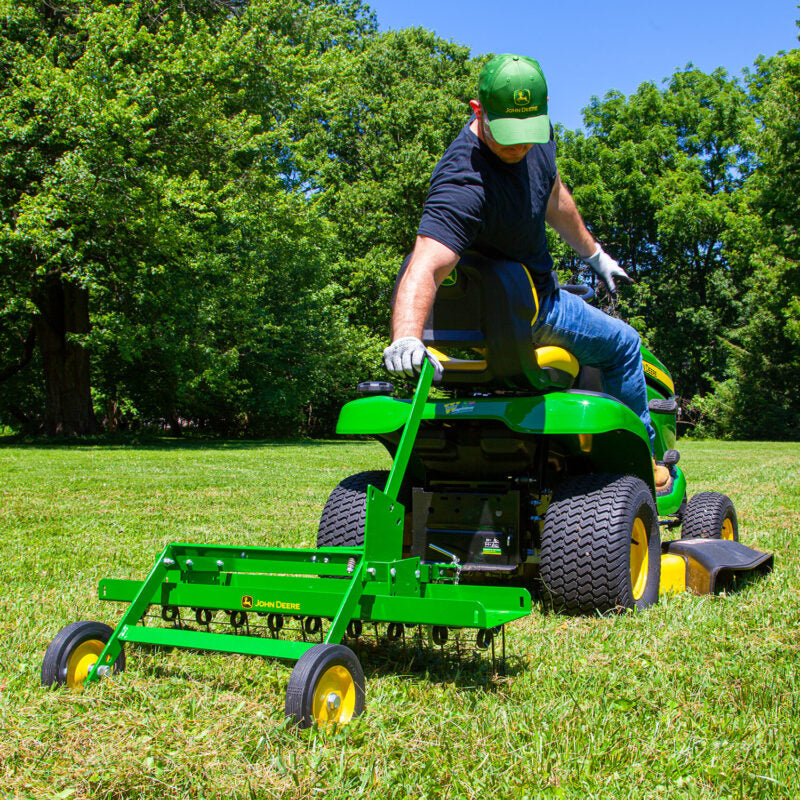 A person in a black shirt and green cap rides a green lawn tractor pulling a green aerator attachment across a grassy field. Trees are visible in the background under a clear blue sky.