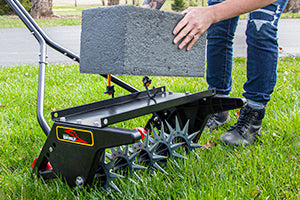A person places a cinder block onto a lawn aerator tool. The aerator has multiple spikes and is sitting on grass. The person is wearing denim pants and black boots.