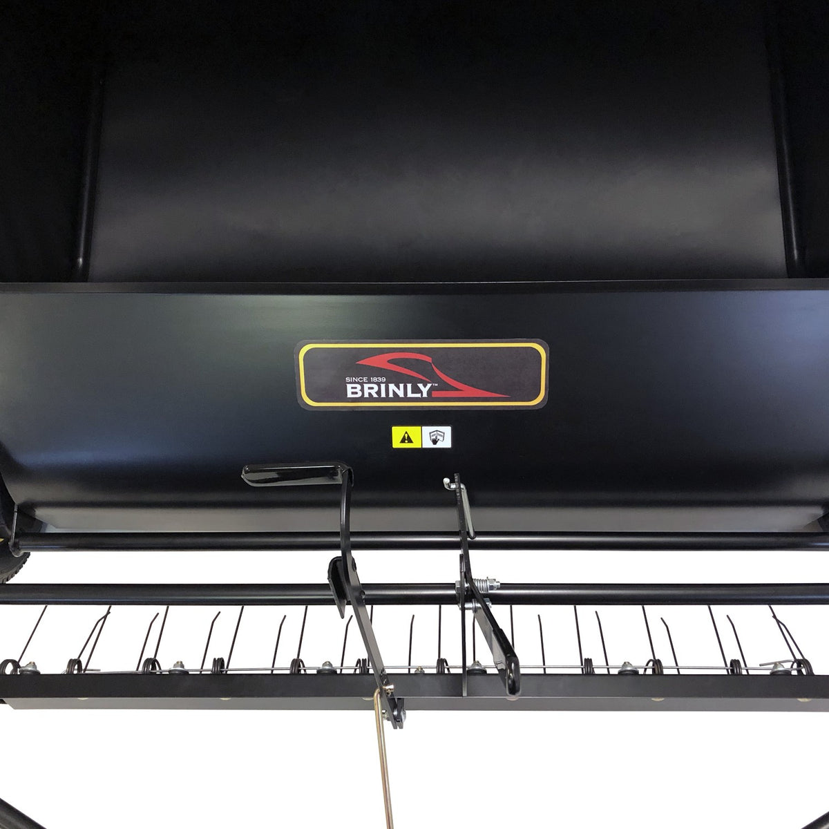 Close-up of a black Brinly Parts 42″ Lawn Sweeper with Dethatcher (STS-42BHDK-A), featuring the company logo, warning label, and portions of the metal frame and sweeper brush assembly.