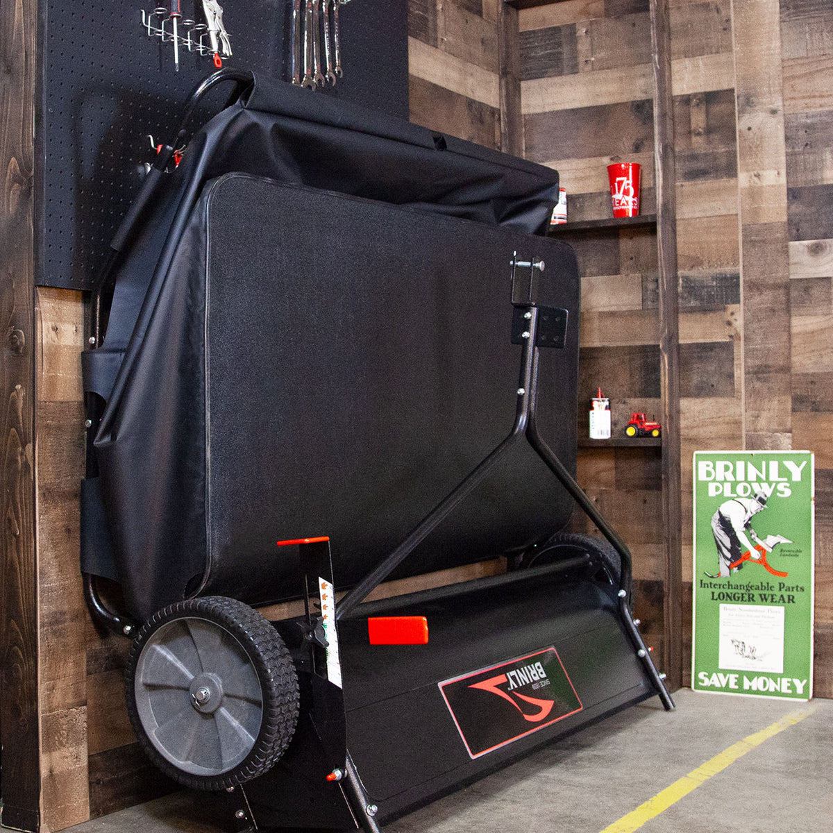 In a garage, a Brinly Parts 54″ Lawn Sweeper with Double-Helix Brushes (LS2-54BH-G) stands against the wooden wall. Nearby, theres a sign reading BRINLY PLOWS with a green tractor image, while tools and small red cans suggest an organization as precise as the sweepers double-helix brush design.