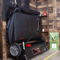In a garage, a Brinly Parts 54″ Lawn Sweeper with Double-Helix Brushes (LS2-54BH-G) stands against the wooden wall. Nearby, theres a sign reading BRINLY PLOWS with a green tractor image, while tools and small red cans suggest an organization as precise as the sweepers double-helix brush design.