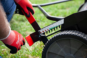 Close-up of a person wearing red gloves adjusting the height lever on a lawn mower. The lever is next to a large wheel on a patch of green grass, showing a scale indicating different height settings.