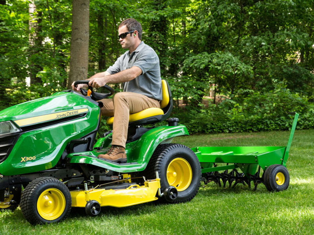 A person wearing sunglasses drives a green John Deere X300 riding mower, towing a small green cart, on a lush lawn surrounded by trees. The mower is in motion, cutting the grass.