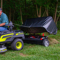 Using a black riding mower with neon green details, someone tows a Brinly Parts 54″ Lawn Sweeper (LS2-54BH-G) filled with grass clippings. Near the woods, the sweeper, featuring a double-helix brush and black tarp, efficiently collects every stray blade.