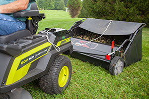 A person operates a black and green riding lawn mower with an attached grass sweeper. The sweeper has a cover partially open, revealing collected grass. The background features green grass and trees.