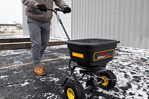 Person pushing a black wheeled salt spreader on a snowy surface next to a building. The spreader has yellow details and a handle. The person is wearing a gray jacket, gray pants, gloves, and brown boots.