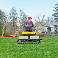 A person in a red cap and plaid shirt rides a yellow lawn mower across a green lawn while effortlessly towing the Brinly Parts 40 Tow-Behind Spike Aerator (SAT2-40BH-P) under a clear sky, with trees and a house in the background ensuring ideal conditions for effective lawn aeration.