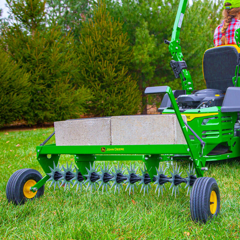 A green lawn aerator attached to a John Deere tractor is being used on a grassy lawn. The aerator has cinder blocks on top for added weight. In the background, there are green trees and a person partially visible on the tractor.