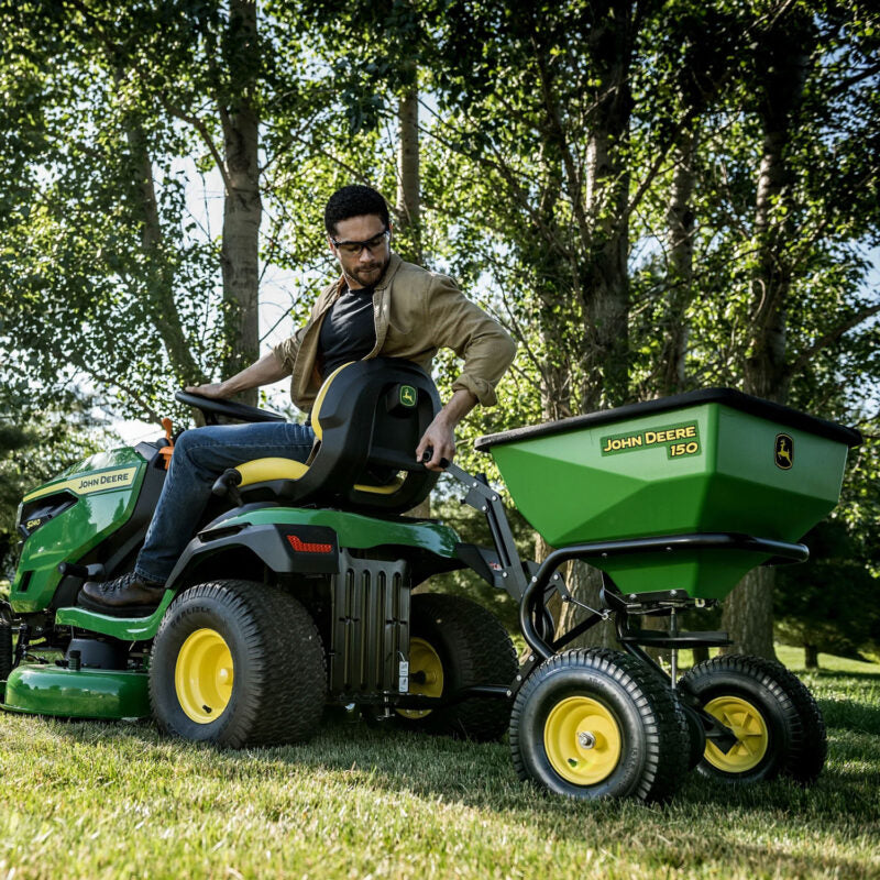 A person is driving a green John Deere lawn tractor with a matching trailer attached. The person is seated, holding the steering wheel, and is wearing a brown jacket and jeans. Trees and grass are in the background.