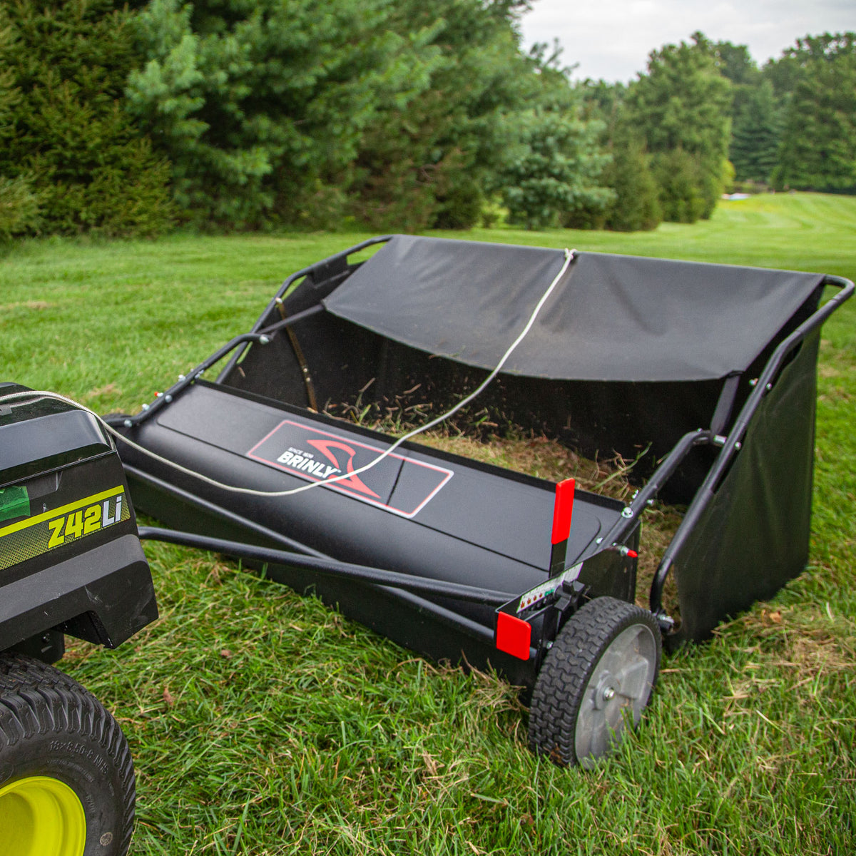 The Brinly Parts 54″ Lawn Sweeper with Double-Helix Brushes, LS2-54BH-G model, efficiently collects grass clippings on a field, maintaining pristine lawns against an overcast sky.