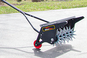 A lawn aerator with a black metal frame, red wheel, and silver spiked drum is placed on a paved surface. The tool is designed for creating holes in the soil to improve air, water, and nutrient absorption.