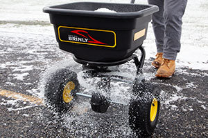 A person pushes a Brinly spreader on a snow-covered path. The spreader dispenses granules onto the ground. The person is wearing gray pants and tan boots. The spreader has yellow wheels and a black container.