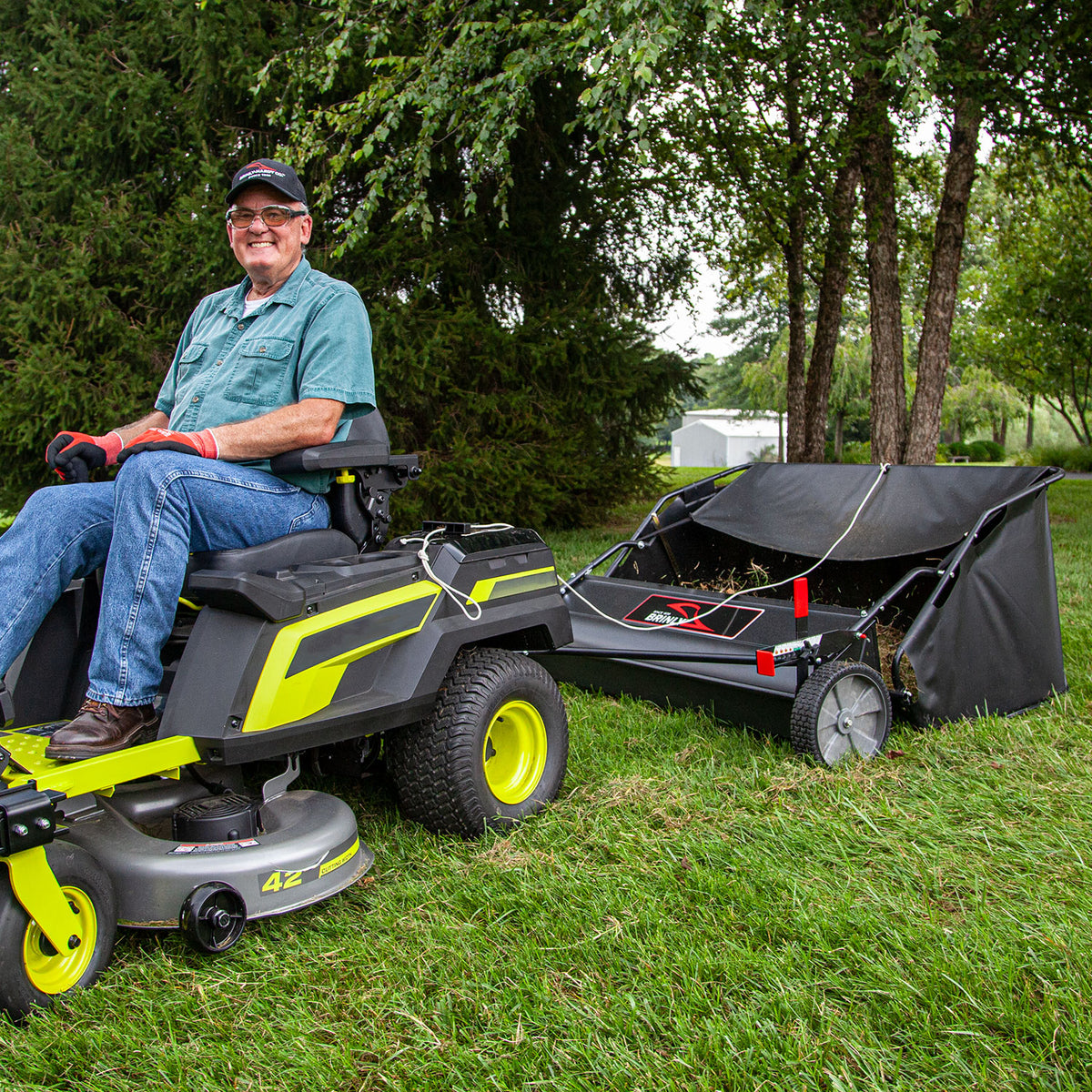 A person in a hat, glasses, gloves, and turquoise shirt smiles while sitting on a lawnmower towing a Brinly Parts 54 Lawn Sweeper with Double-Helix Brushes (LS2-54BH-G) across a grassy area surrounded by trees.