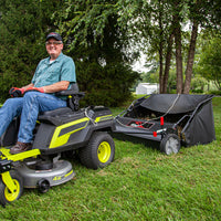 A person in a hat, glasses, gloves, and turquoise shirt smiles while sitting on a lawnmower towing a Brinly Parts 54 Lawn Sweeper with Double-Helix Brushes (LS2-54BH-G) across a grassy area surrounded by trees.