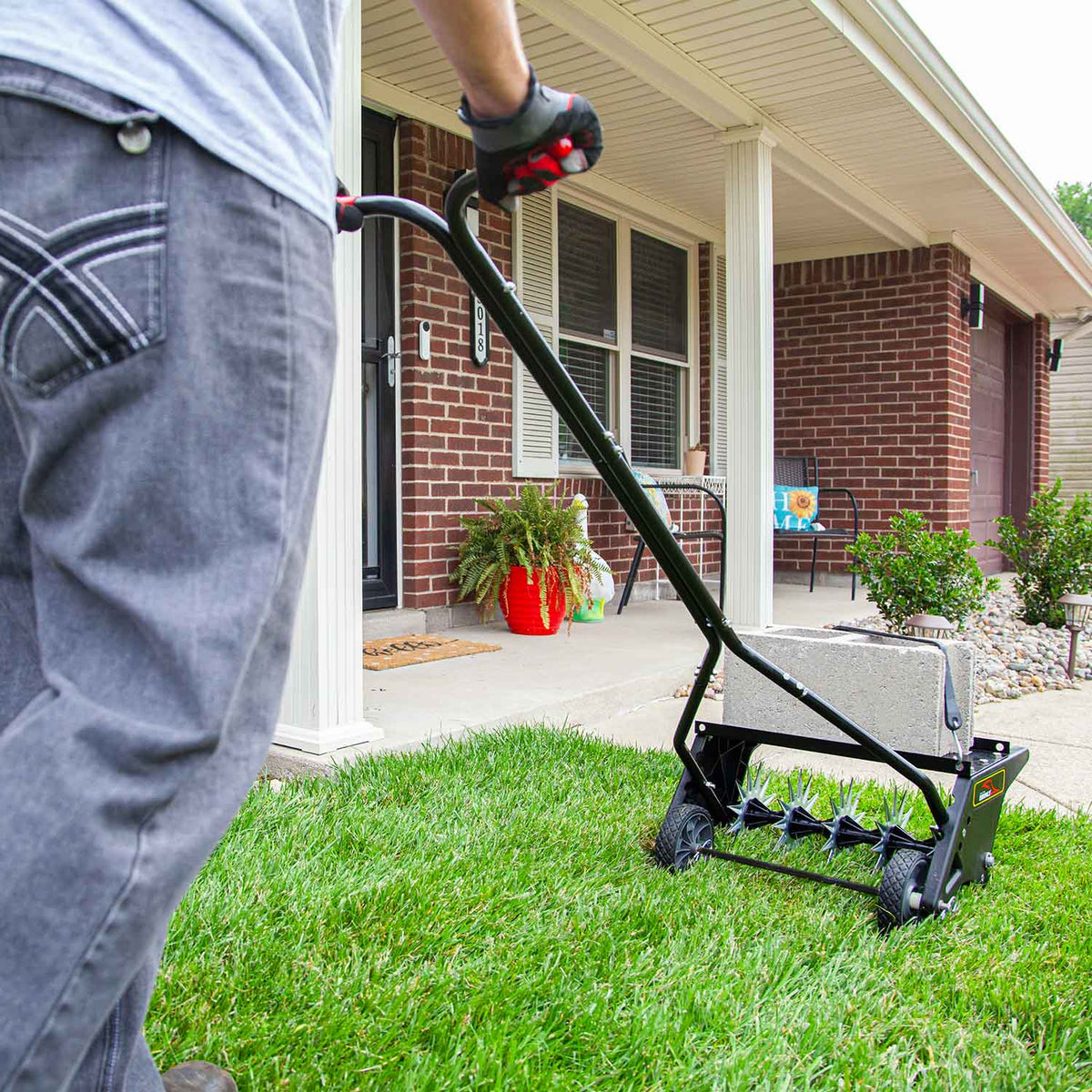 Wearing jeans and gloves, a person uses a manual reel mower to trim the grass in front of a brick house with a porch featuring a red planter and cozy seating. Nearby, the Brinly Parts 20 Push Spike Aerator with 3D Galvanized Steel Tines suggests plans for lawn aeration on this sunny day.