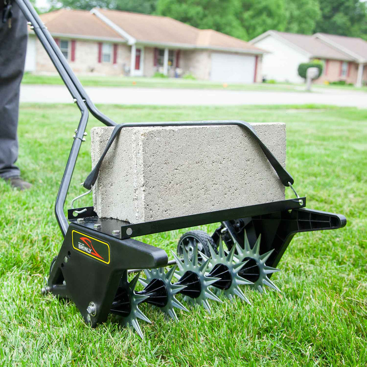 A Brinly Parts 20 Push Spike Aerator with 3D Galvanized Steel Tines is efficiently aerating grass, enhanced by a concrete block for added weight. Houses can be seen in the background.