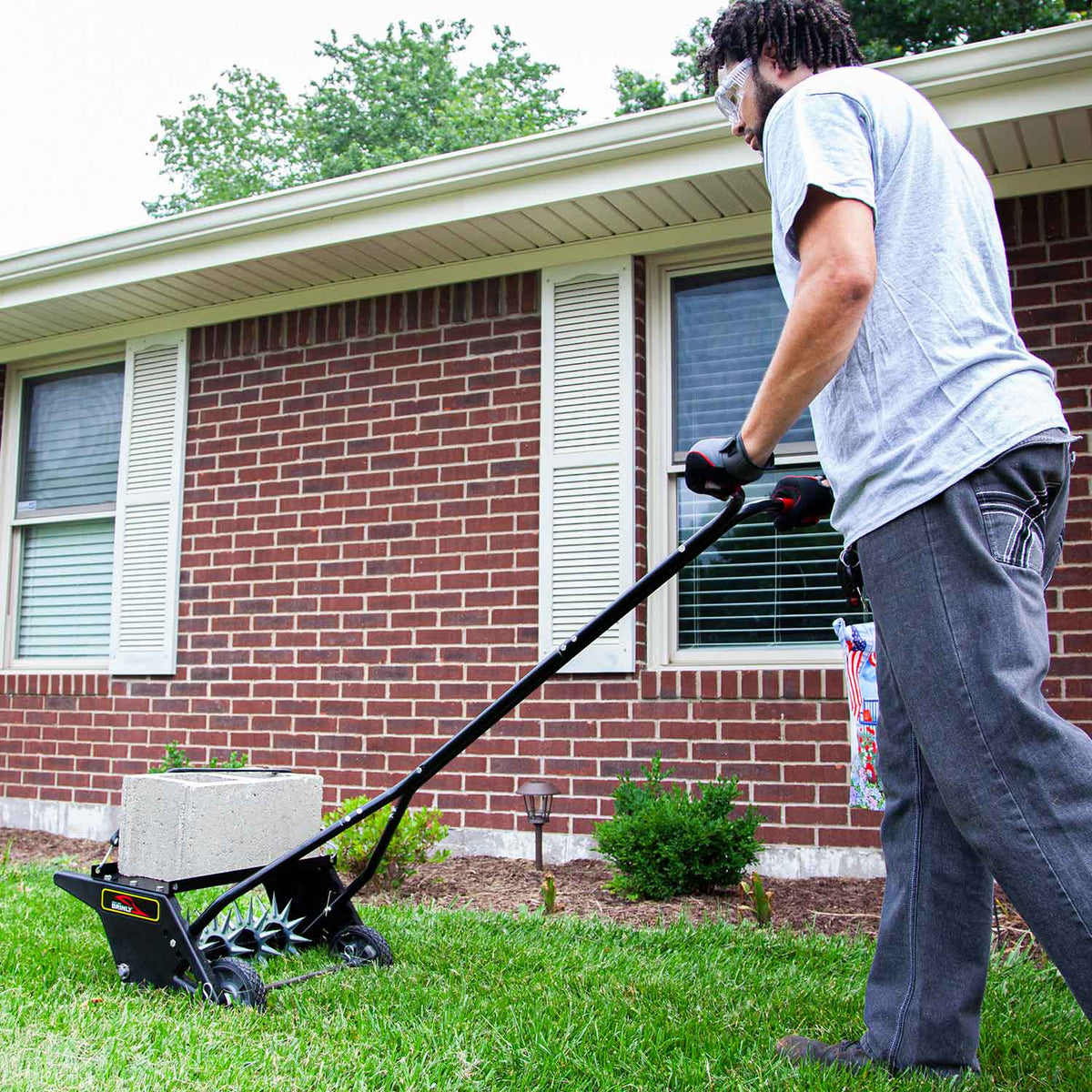 A person mowing a lawn with a manual reel mower in front of a brick house, donned in a gray shirt, jeans, and glasses. The lush green grass suggests they might regularly use a Brinly Parts 20 Push Spike Aerator with 3D Galvanized Steel Tines. A small garden bed sits nearby.