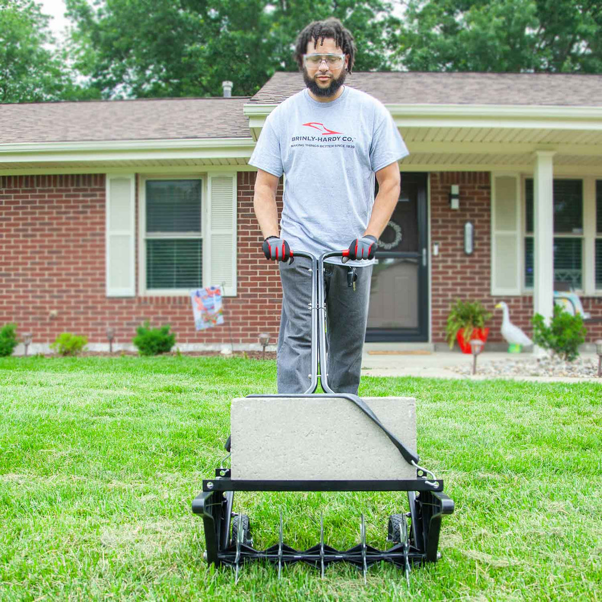 Wearing glasses and gray attire with gloves, a person uses a reel mower to invigorate the lawn before a brick house with white shutters and a porch. Under an overcast sky, they work nearby where a Brinly Parts 20 Push Spike Aerator with 3D Galvanized Steel Tines rests against the wall.