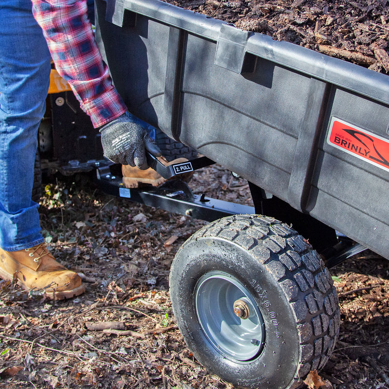 Wearing jeans, a plaid shirt, gloves, and work boots, a person pulls a lever on a Brinly Parts cart. The Roger 180-Degree Full Dump Kit for Brinly-Hardy 10 cu. ft. Poly Carts effortlessly dumps soil or mulch amidst scattered leaves and twigs.