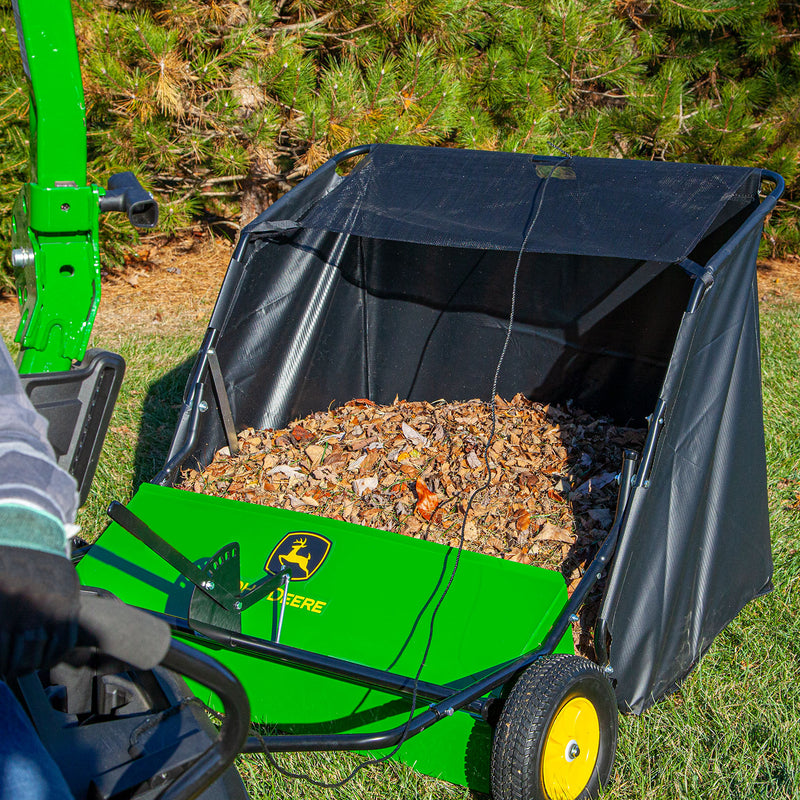 A green John Deere 42 Tow-Behind Lawn Sweeper (LPSTS-42JD / STS-42JD) with a high-capacity hamper, filled with dry leaves and attached to a tractor, sits on a grassy lawn near trees.