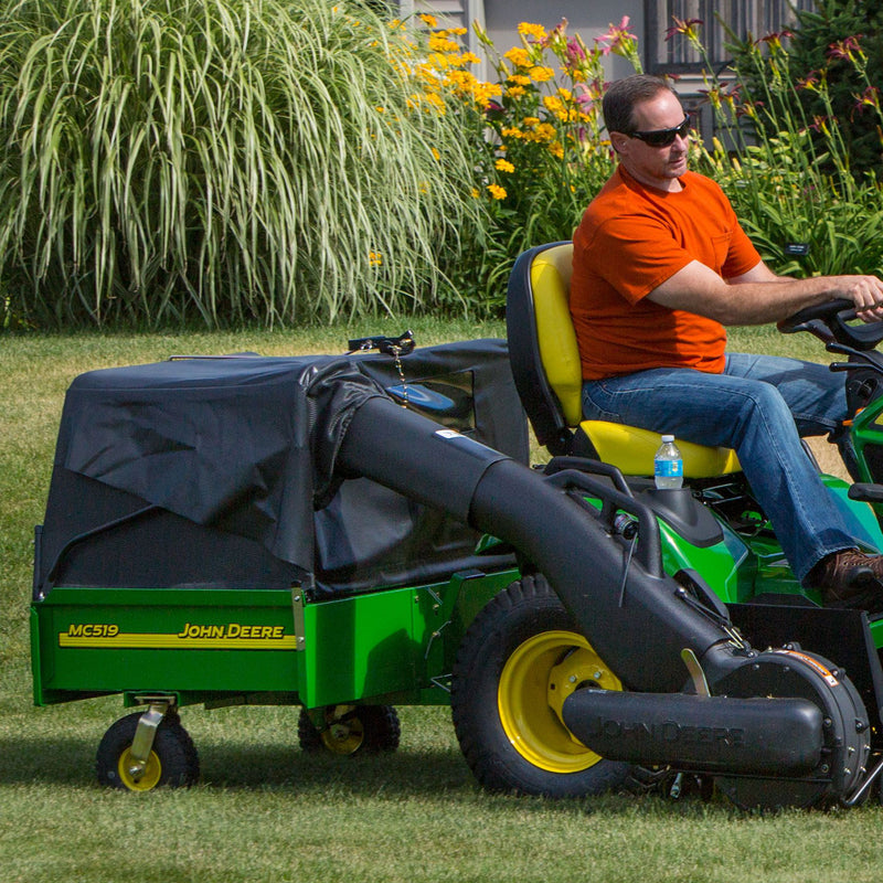 A person in a red shirt and sunglasses rides a green John Deere lawnmower with the John Deere MC519 Material Collection System (LP49228) attached. Tall grass and yellow flowers decorate the garden, while a water bottle sits in the storage area.