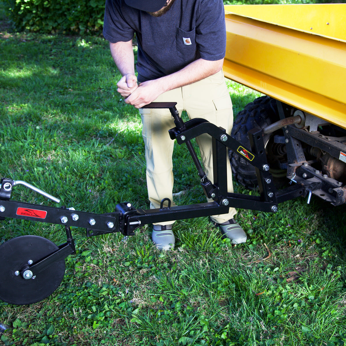 A person in a blue shirt, khaki pants, and sneakers attaches a black Brinly Parts Universal ATV/UTV One-Point Lift (OPL2-BH) for Brinly Ground Engaging Products to a yellow utility vehicle outdoors on grass.
