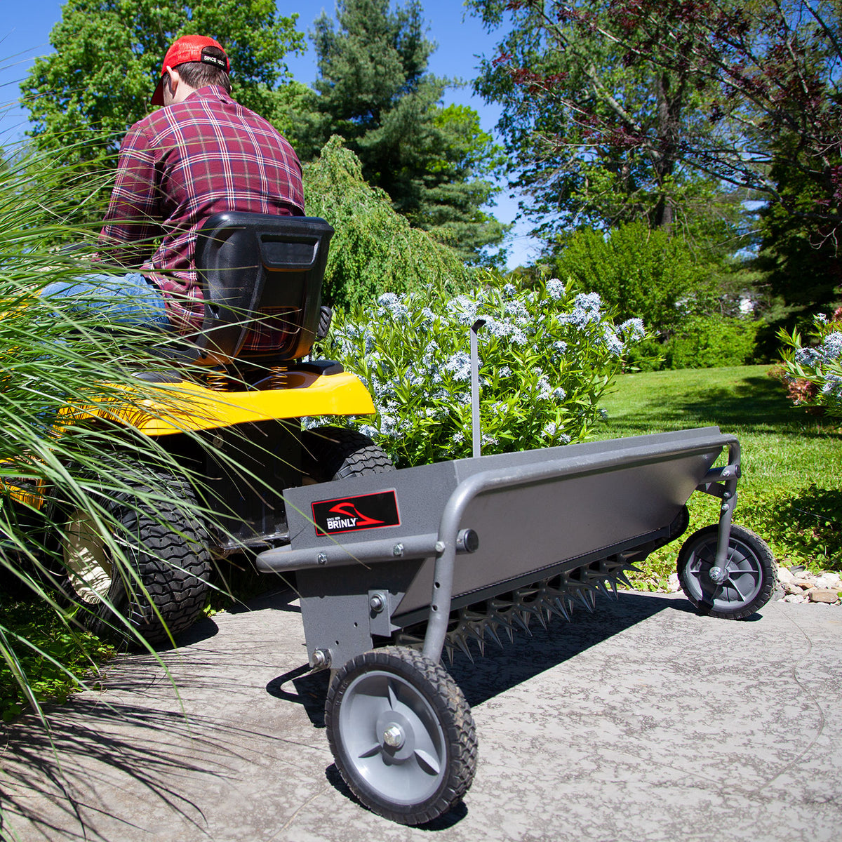 A person in a red cap and plaid shirt rides a yellow lawn tractor, towing a Brinly Parts 40 Combination Aerator Spreader in Hammered Gunmetal on a stone path, surrounded by lush green grass, bushes, and trees under a clear blue sky.