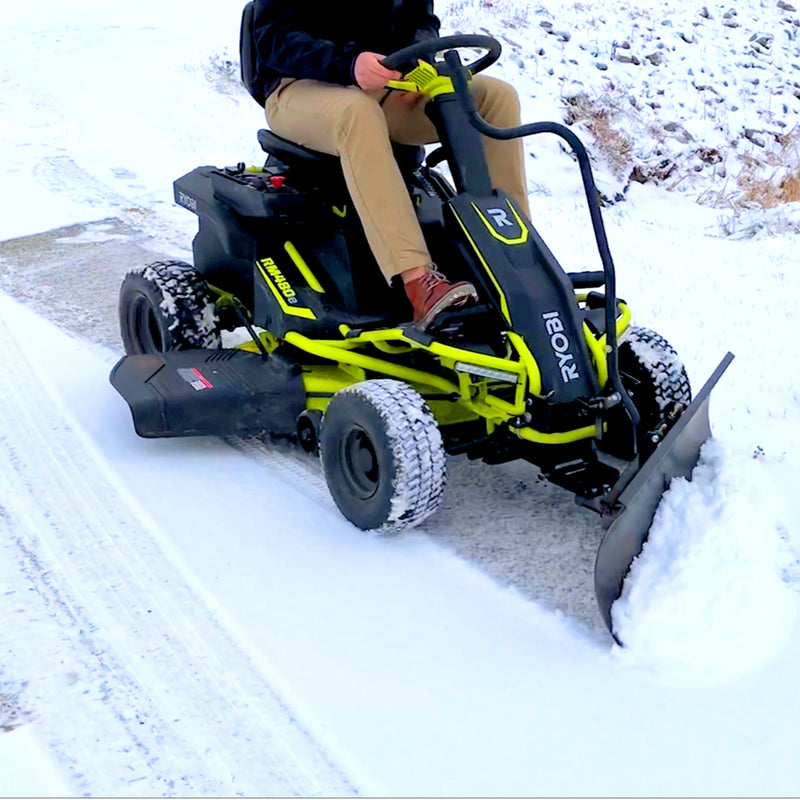 A person operates a Ryobi Electric Riding Mower, fitted with a Brinly Parts 42 in. Front Mount Blade (FB-42RY), easily clearing snow. The vivid green and black scheme contrasts as the steel blade unveils the pavement beneath. Theyre dressed in beige pants, focused on their task.